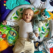 Jahlënn a rejoint le concours — aidez-le/la à gagner de superbes lots ! baby, child, infant, play_mat, soft_toys, book, white_shirt, beige_pants, floor_tiles, colorful, smiling, cute, young_child, indoor, happy, lying_down, stuffed_animal, mirror, leaf_pattern, toy