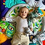 baby, child, infant, play_mat, soft_toys, book, white_shirt, beige_pants, floor_tiles, colorful, smiling, cute, young_child, indoor, happy, lying_down, stuffed_animal, mirror, leaf_pattern, toy
