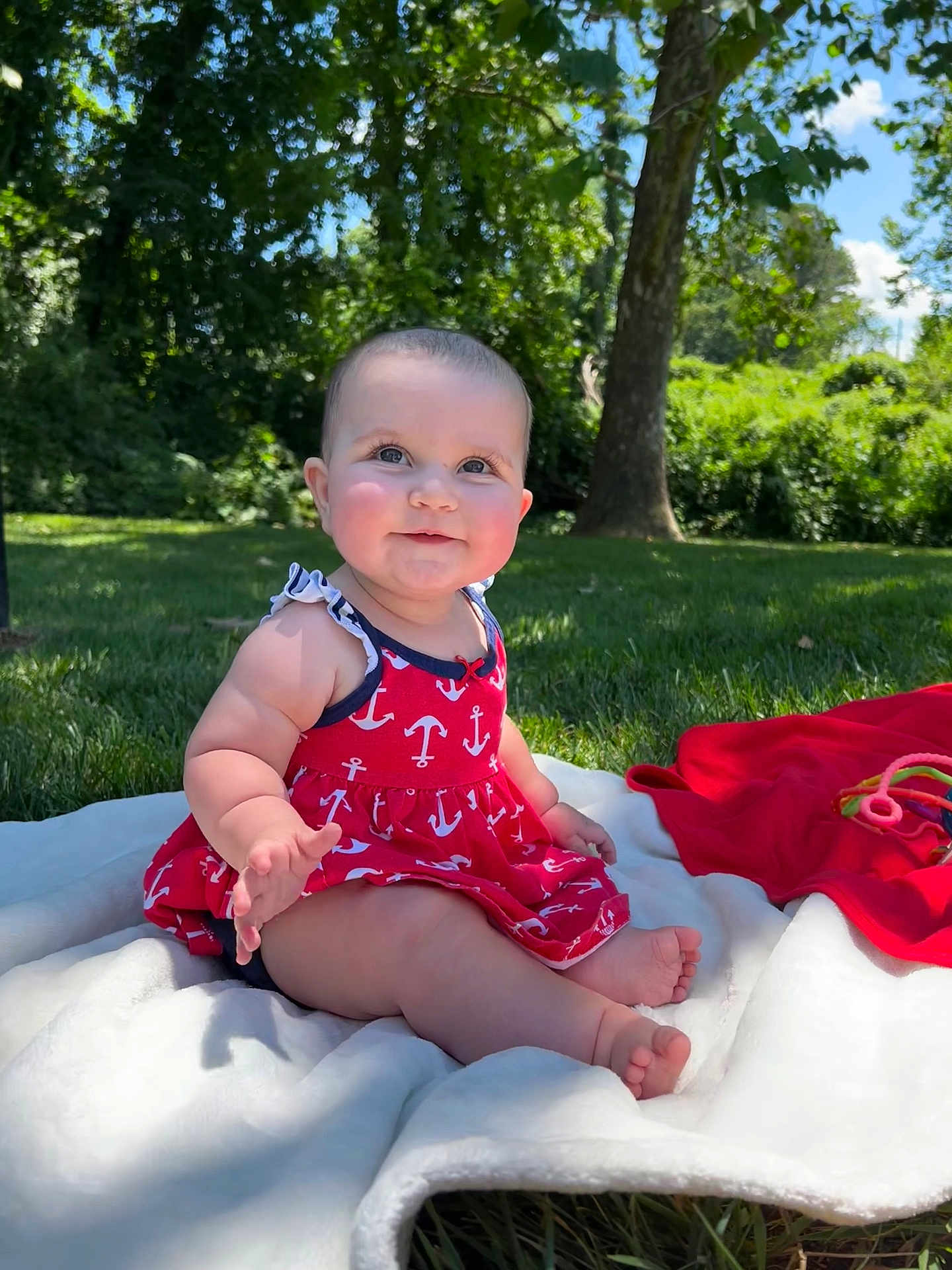 Iris is registered to the contest to win money with this photo: baby, child, outdoor, grass, tree, blanket, dress, red, anchor_pattern, smiling, sitting, cute, sunlight, nature, playtime, barefoot, happy, person, infant, summer
