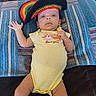 baby, infant, yellow_onesie, crocheted_hat, striped_towel, blanket, lying_down, face, headwear, clothing, person, child, cute, indoors, skin, hand, legs, expression, colorful, soft_surface