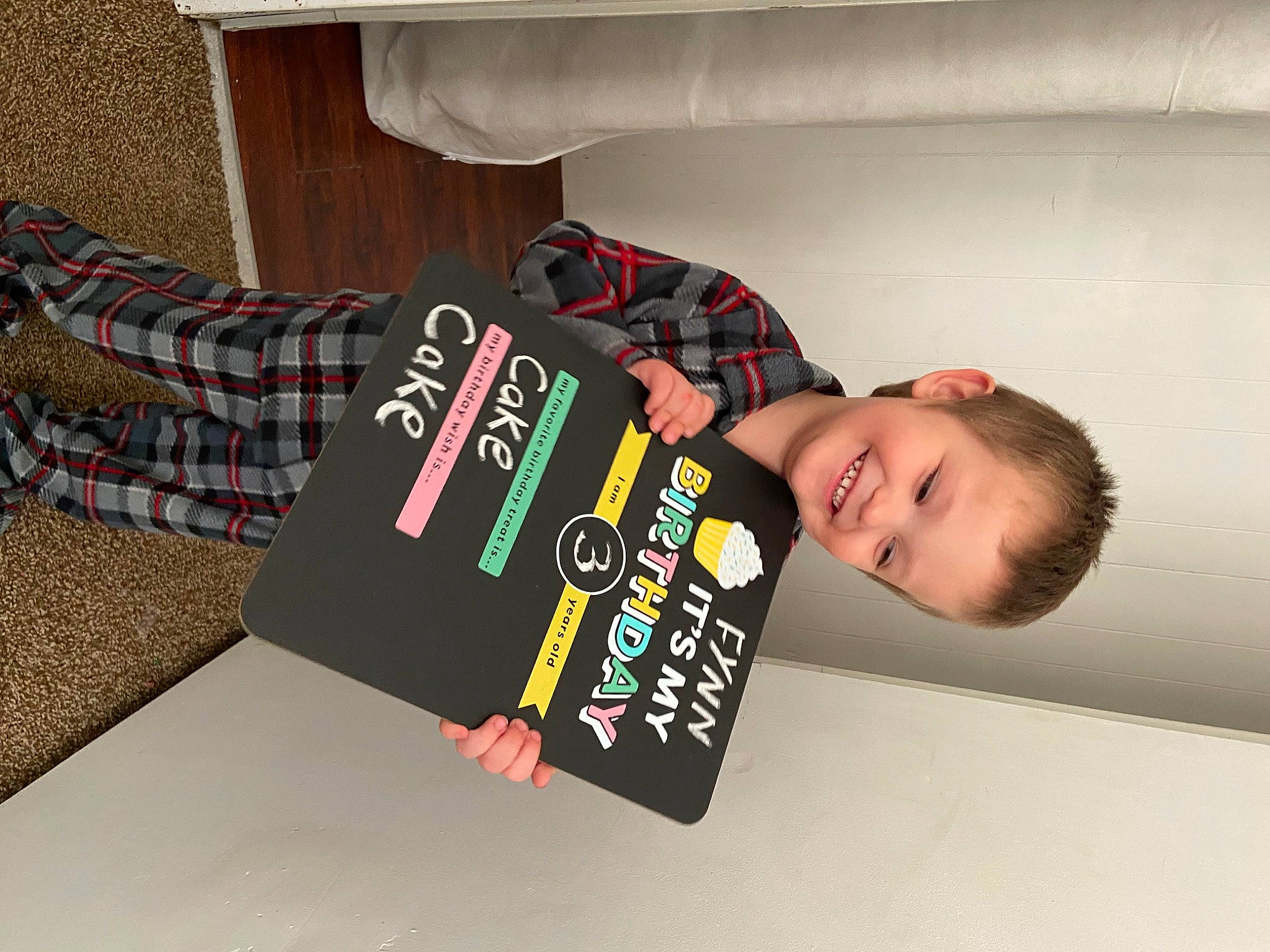 Fynn is registered to the contest to win money with this photo: child, comfort, elbow, flooring, fun, happy, joy, pattern, person, plaid, room, sleeve, smile, t_shirt, table, tartan, textile, toddler, top, wrist