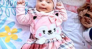 Kylie is registered to the contest to win money with this photo: baby, infant, pink_clothing, plaid, dress, hat, smiling, cute, lying_down, blanket, floral_pattern, footwear, indoors, child, person, face, headwear, happy, soft_toys, cozy