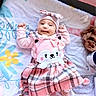 baby, infant, pink_clothing, plaid, dress, hat, smiling, cute, lying_down, blanket, floral_pattern, footwear, indoors, child, person, face, headwear, happy, soft_toys, cozy