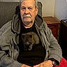 Church is registered to the contest to win money with this photo: man, elderly, kitten, cat, pet, portrait, indoors, armchair, sitting, gray_hoodie, black_shirt, red_blanket, living_room, furniture, window, blinds, wooden_table, watch, hands, cozy