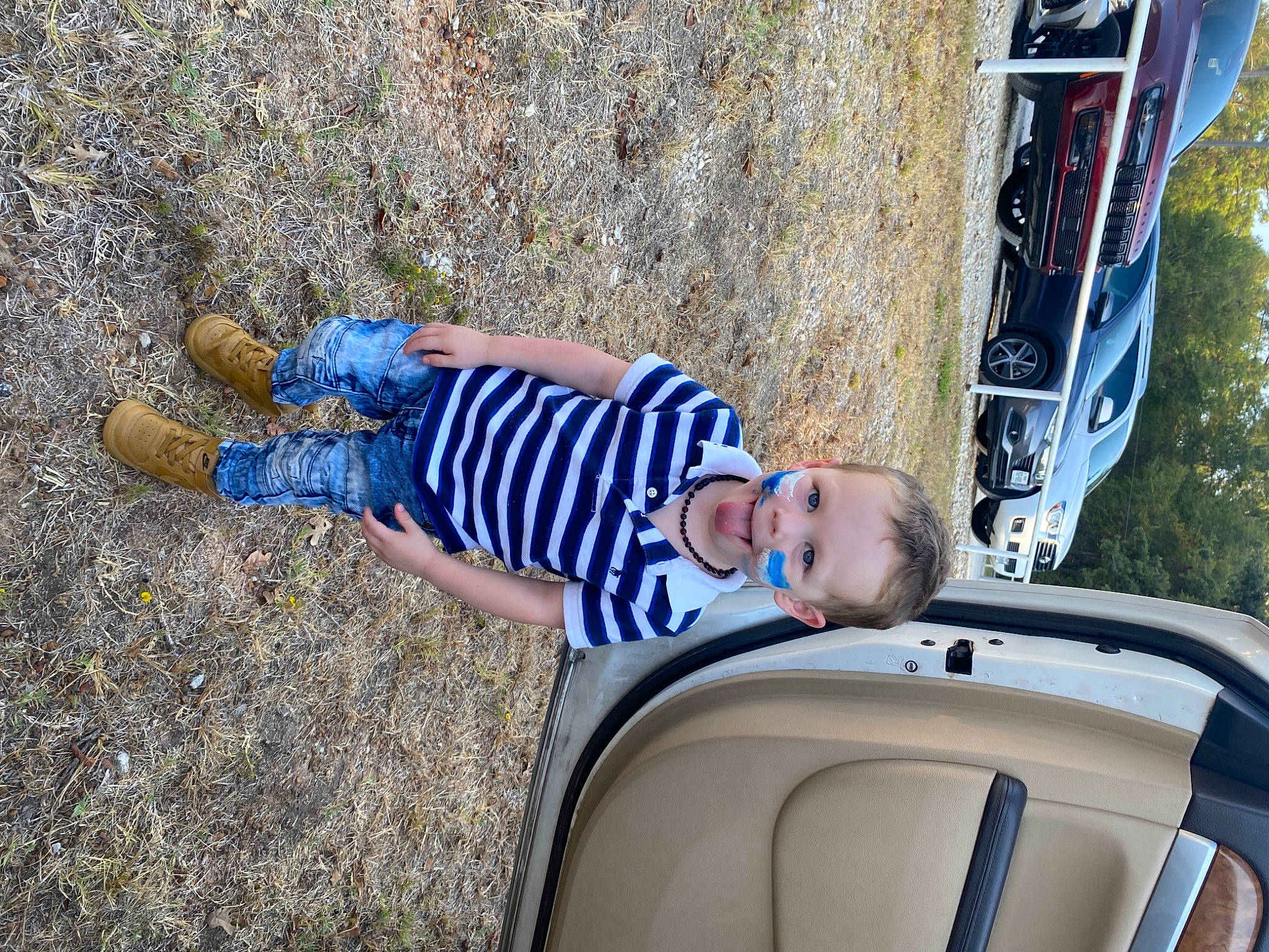 Bentley is registered to the contest to win money with this photo: asphalt, auto_part, automotive_exterior, automotive_tire, car, child, electric_blue, family_car, fun, grass, hood, human_leg, leisure, motor_vehicle, person, recreation, sitting, sunglasses, toddler, tree