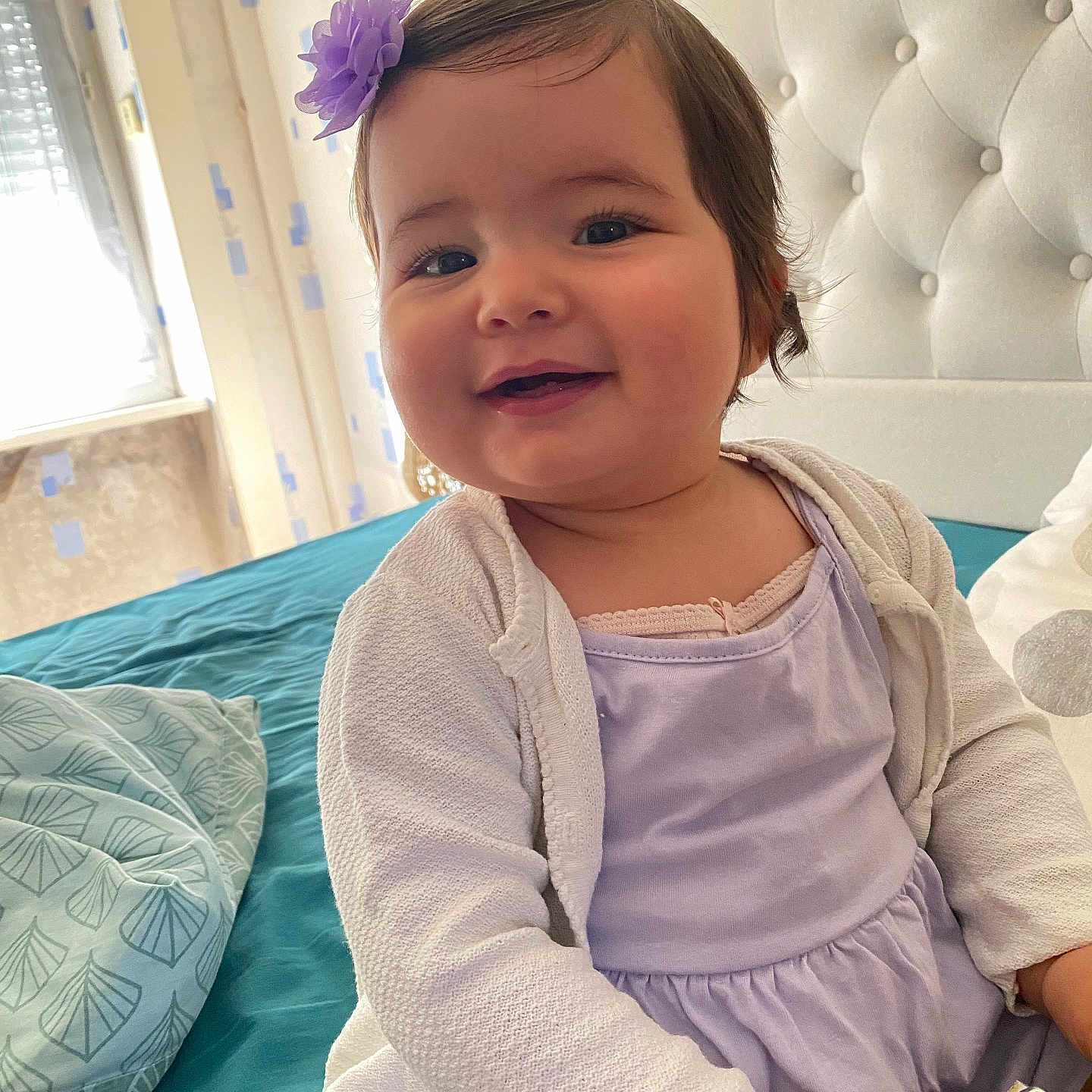 Giulia participe au concours pour gagner de l'argent avec cette photo : baby, bed, child, cute, flower_clip, girl, happy, headboard, indoor, infant, natural_light, person, pillow, portrait, purple_dress, smiling, teal_bedspread, wallpaper, white_cardigan, window