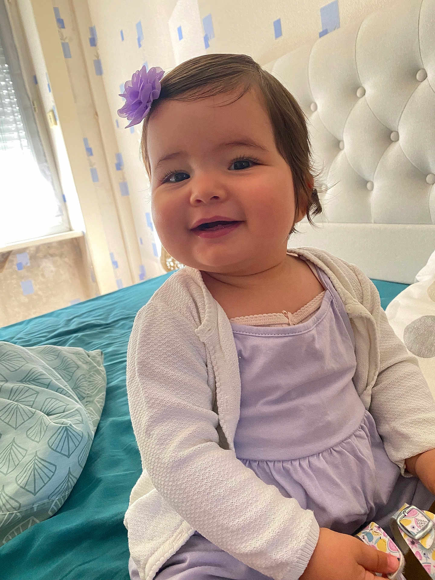 Giulia participe au concours pour gagner de l'argent avec cette photo : baby, child, smiling, girl, flower_clip, purple_dress, white_cardigan, bed, pillow, teal_bedspread, indoor, portrait, happy, cute, headboard, wallpaper, window, natural_light, person, infant