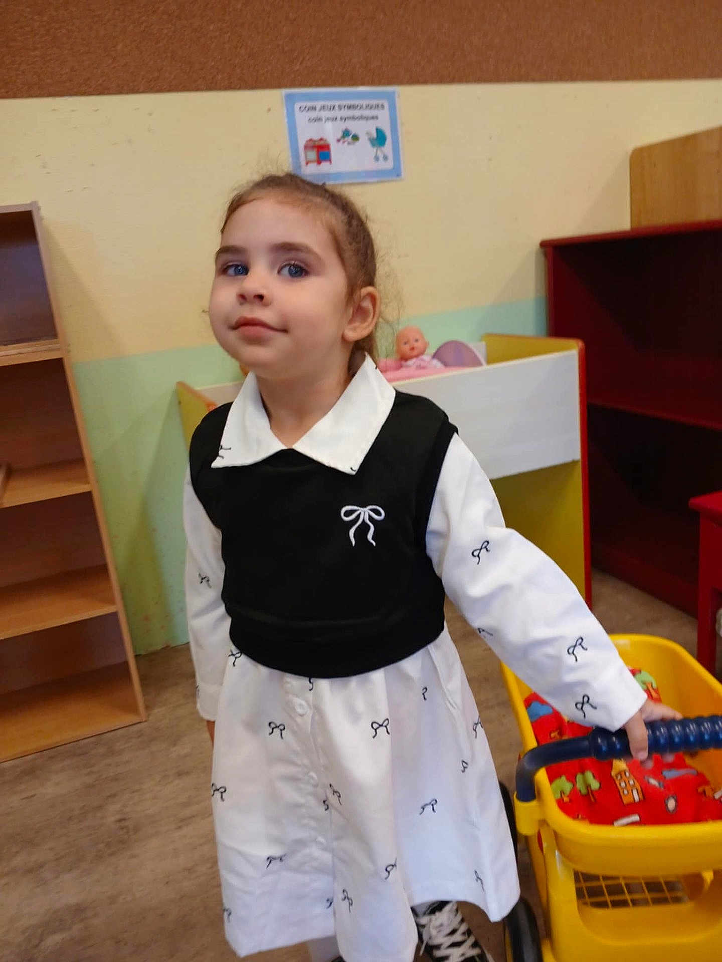 Ilona a rejoint le concours — aidez-le/la à gagner de superbes lots ! child, girl, toy_cart, doll, playroom, white_dress, black_vest, bow_pattern, floor, shelf, wall, toy, indoor, person, young_child, standing, smile, casual, shoes, colorful