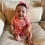 Sofia is registered to the contest to win money with this photo: baby, child, smile, pink_headband, striped_dress, couch, cushion, indoor, happy, sitting, cute, infant, small_toes, hand, portrait, person, comfortable, soft_furniture, home, bright