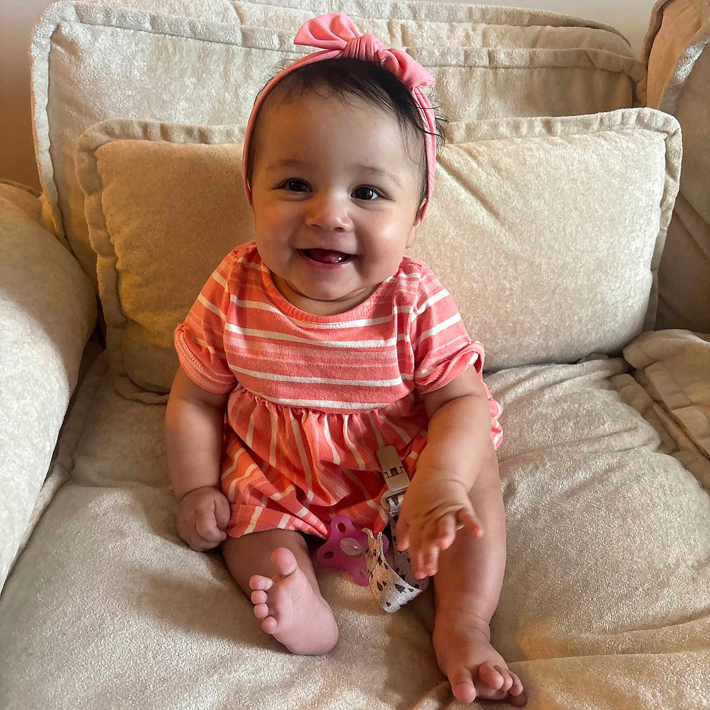 Sofia is registered to the contest to win money with this photo: baby, child, smile, pink_headband, striped_dress, couch, cushion, indoor, happy, sitting, cute, infant, small_toes, hand, portrait, person, comfortable, soft_furniture, home, bright