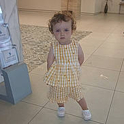 Emily joined the competition — help win amazing prizes! toddler, child, yellow_dress, curly_hair, indoor, tile_floor, serious_expression, person, shoes, standing, lobby, plant, reception_desk, carpet, furniture, pot, woman, desk, light, casual_clothing