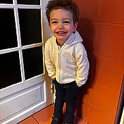 Ilyes a rejoint le concours — aidez-le/la à gagner de superbes lots ! child, toddler, smile, curly_hair, hoodie, white_hoodie, jeans, black_shoes, door, tiles, orange_wall, standing, indoor, happy, portrait, hands_in_pockets, floor_tiles, grin, person, young_child