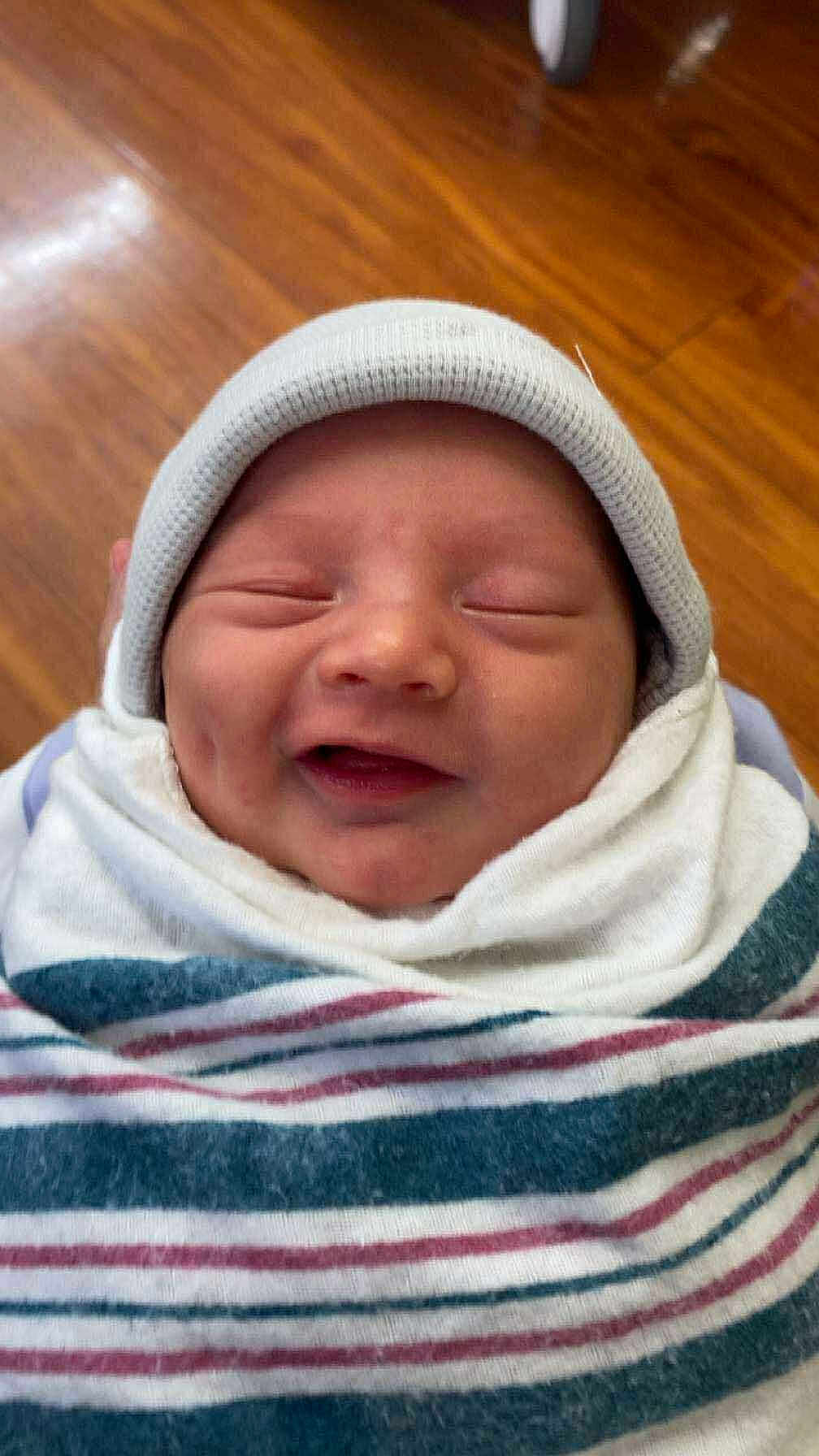 Ashton is registered to the contest to win money with this photo: baby, newborn, smiling, hat, blanket, striped_blanket, cozy, sleepy, face, infant, child, wrapped, cute, person, headwear, happy, indoors, wooden_floor, portrait, resting