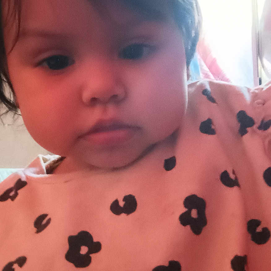Rosemarie is registered to the contest to win money with this photo: baby, black_flower_pattern, casual_clothing, child, closeup, curtain, expressionless, face, hair, head, home_interior, indoor, lamp, person, pink_shirt, portrait, soft_lighting, toddler, window, young_child