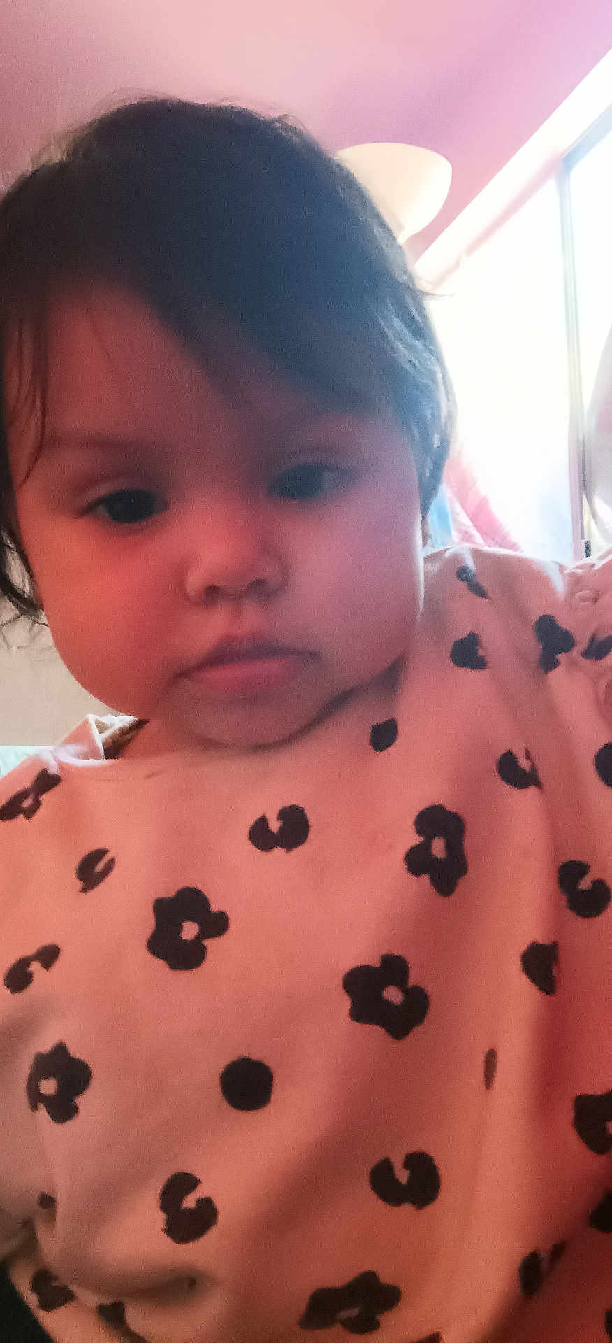 Rosemarie is registered to the contest to win money with this photo: child, toddler, face, indoor, pink_shirt, black_flower_pattern, hair, portrait, expressionless, young_child, person, window, lamp, soft_lighting, closeup, casual_clothing, home_interior, curtain, baby, head