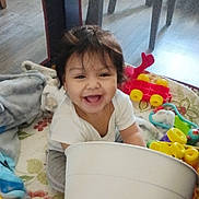 Rosemarie is registered to the contest to win money with this photo: toddler, child, smiling, playpen, toys, bucket, blanket, indoor, happy, baby, floor, plastic_toy, clothing, person, home, colorful, sitting, cute, young_child, playtime