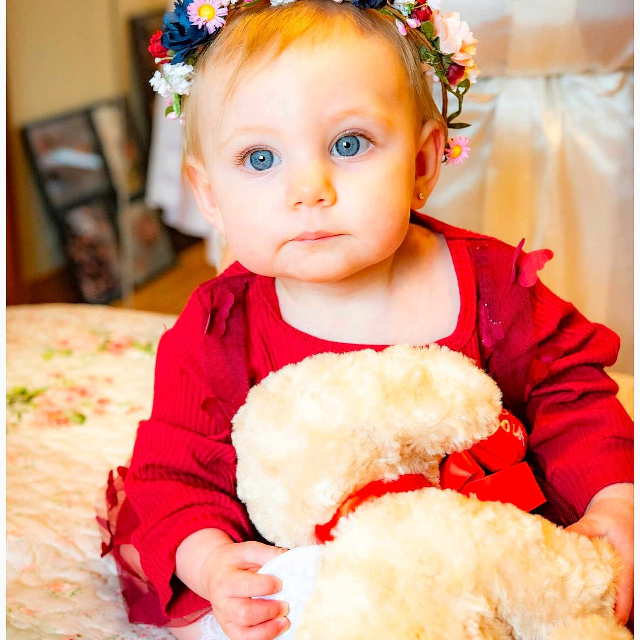 Sophie is registered to the contest to win money with this photo: baby, bedspread, blue_eyes, child, cute, decor, face, flower_crown, hands, indoor, infant, person, plush_toy, portrait, red_dress, sitting, soft_lighting, teddy_bear, warm_colors, young_child