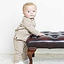toddler, child, infant, pajamas, beige_clothing, leather_ottoman, tufted_furniture, white_background, carpet, standing, looking_at_camera, indoor, cute, baby, portrait, small_child, furniture, hand_on_furniture, floor, socks