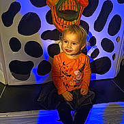 Tallulah is registered to the contest to win money with this photo: child, toddler, halloween, orange_shirt, peppa_pig, skull_mask, spooky, blue_light, smiling, sitting, indoor, costume, black_tights, decor, face, cute, festive, holiday, background, playful