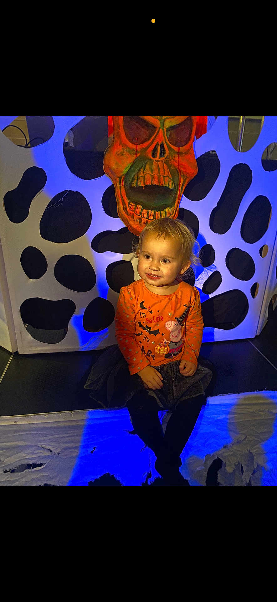Tallulah is registered to the contest to win money with this photo: child, toddler, halloween, orange_shirt, peppa_pig, skull_mask, spooky, blue_light, smiling, sitting, indoor, costume, black_tights, decor, face, cute, festive, holiday, background, playful