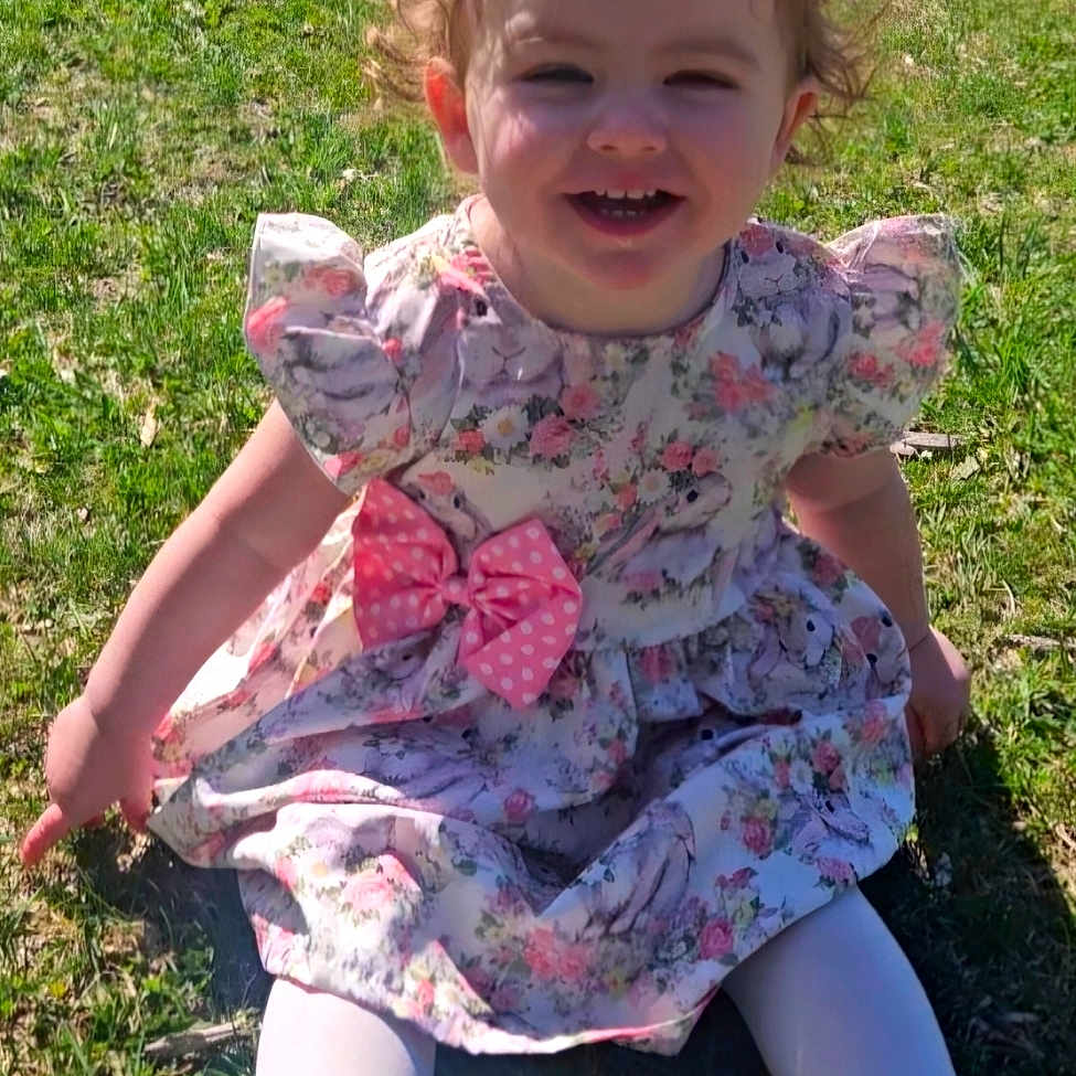 Michelle Shelly joined the competition — help win amazing prizes! bow, child, curly_hair, cute, dress, flower_pattern, girl, grass, happy, nature, outdoor, pink, playful, portrait, sitting, smiling, sunlight, toddler, white_shoes, white_tights