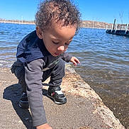 Nazir is registered to the contest to win money with this photo: child, toddler, curly_hair, hoodie, sneakers, lake, water, sky, outdoor, nature, sand, shore, dock, sunny, daytime, person, exploring, crouching, curiosity, casual_clothing