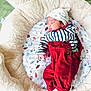 newborn, baby, sleeping, plush_toy, teddy_bear, bed, red_overalls, striped_shirt, hat, cushion, leaf_pattern, green_blanket, infant, cozy, soft, cute, child, resting, indoors, comfort