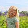 child, girl, smiling, blonde_hair, flower_field, purple_flowers, greenery, outdoor, nature, sky, clouds, summer, casual_clothing, short_sleeve, happy, portrait, grass, sunlight, playful, scenic