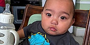 Anthony is registered to the contest to win money with this photo: baby, bottle, child, clothing, cup, drink, expression, face, furniture, high_chair, indoor, kitchen, magnet, person, refrigerator, seat, sippy_cup, stove, table, toddler