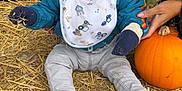 Freddie is registered to the contest to win money with this photo: autumn, bib, blue_jacket, child, curious, fall, gray_pants, hand, hat, mittens, orange, outdoor, person, pumpkin, seasonal, sitting, sneakers, straw, toddler, wide_eyes
