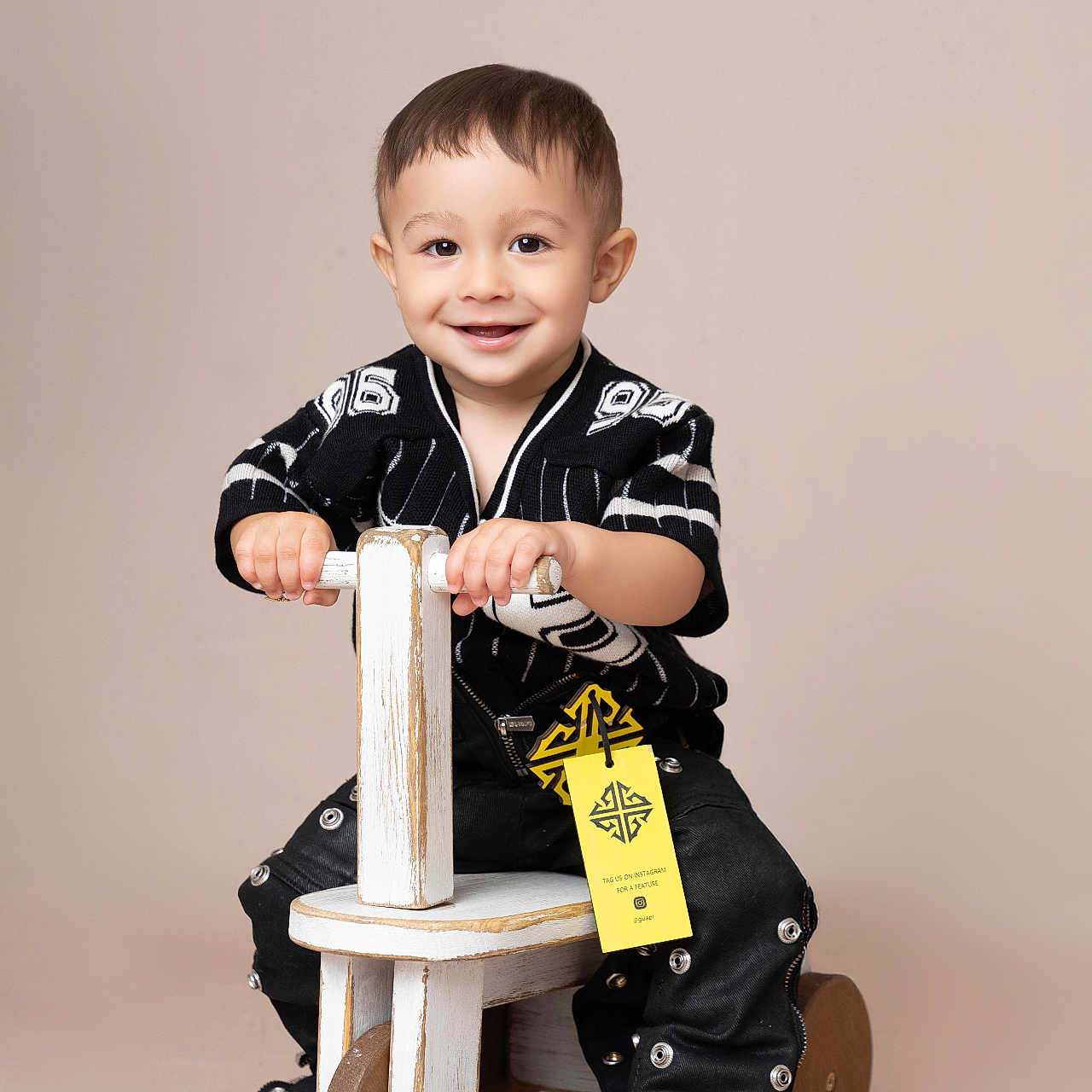 Ean is registered to the contest to win money with this photo: beige_background, black_clothing, child, cute, face, fashion, hands, happy, indoor, pants, play, portrait, ride_on_toy, shoes, short_hair, sitting, smiling, studio_photo, toddler, wooden_toy