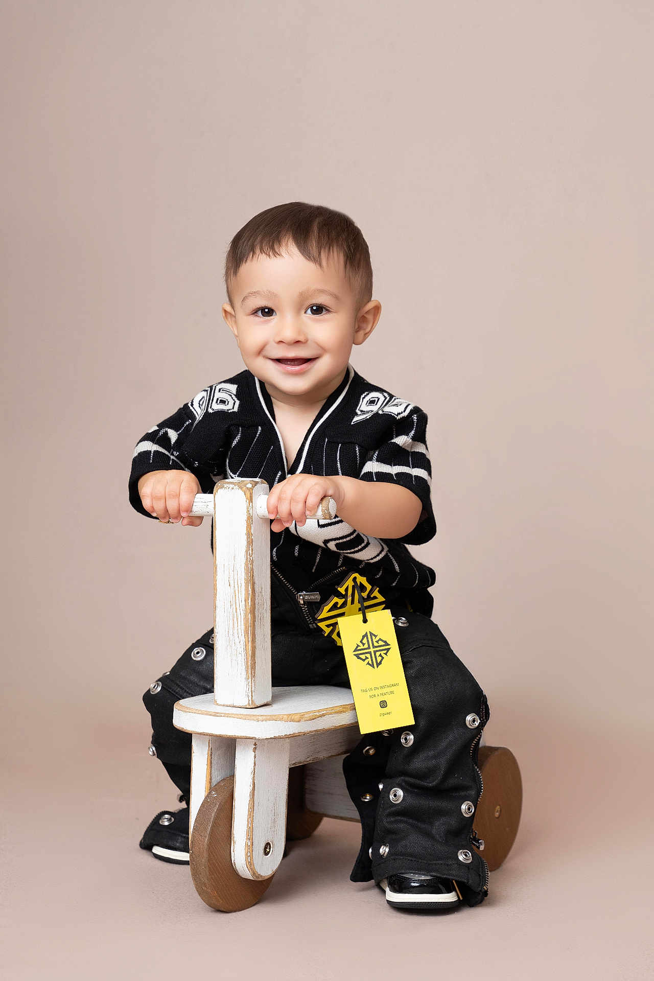 Ean is registered to the contest to win money with this photo: toddler, child, smiling, ride_on_toy, wooden_toy, black_clothing, pants, shoes, studio_photo, beige_background, happy, face, hands, short_hair, sitting, cute, fashion, portrait, indoor, play