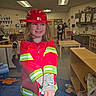 child, firefighter_costume, red_helmet, classroom, toy, table, chair, carpet, person, indoor, play, costume, educational, furniture, smile, childhood, learning, school, kids, expression