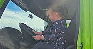 Trinity is registered to the contest to win money with this photo: child, girl, vehicle, steering_wheel, seat, window, sunlight, lens_flare, pony_tail, casual_clothing, sneakers, blue_sky, indoor, side_view, green_color, hands, profile, daylight, driving, seat_belt