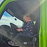 child, girl, vehicle, steering_wheel, seat, window, sunlight, lens_flare, pony_tail, casual_clothing, sneakers, blue_sky, indoor, side_view, green_color, hands, profile, daylight, driving, seat_belt