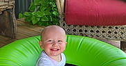 Zaidyn is registered to the contest to win money with this photo: baby, casual_clothing, chair, child, cushion, face, green, happy, head, inflatable_ring, leafy_plant, outdoor, person, plant, porch, sitting, smiling, sunlight, toddler, wood_floor
