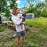 baby, child, cloud, daylight, face, fence, footwear, grass, greenery, holding, hoodie, interaction, nature, outdoor, person, rocks, shed, shorts, sky, tree