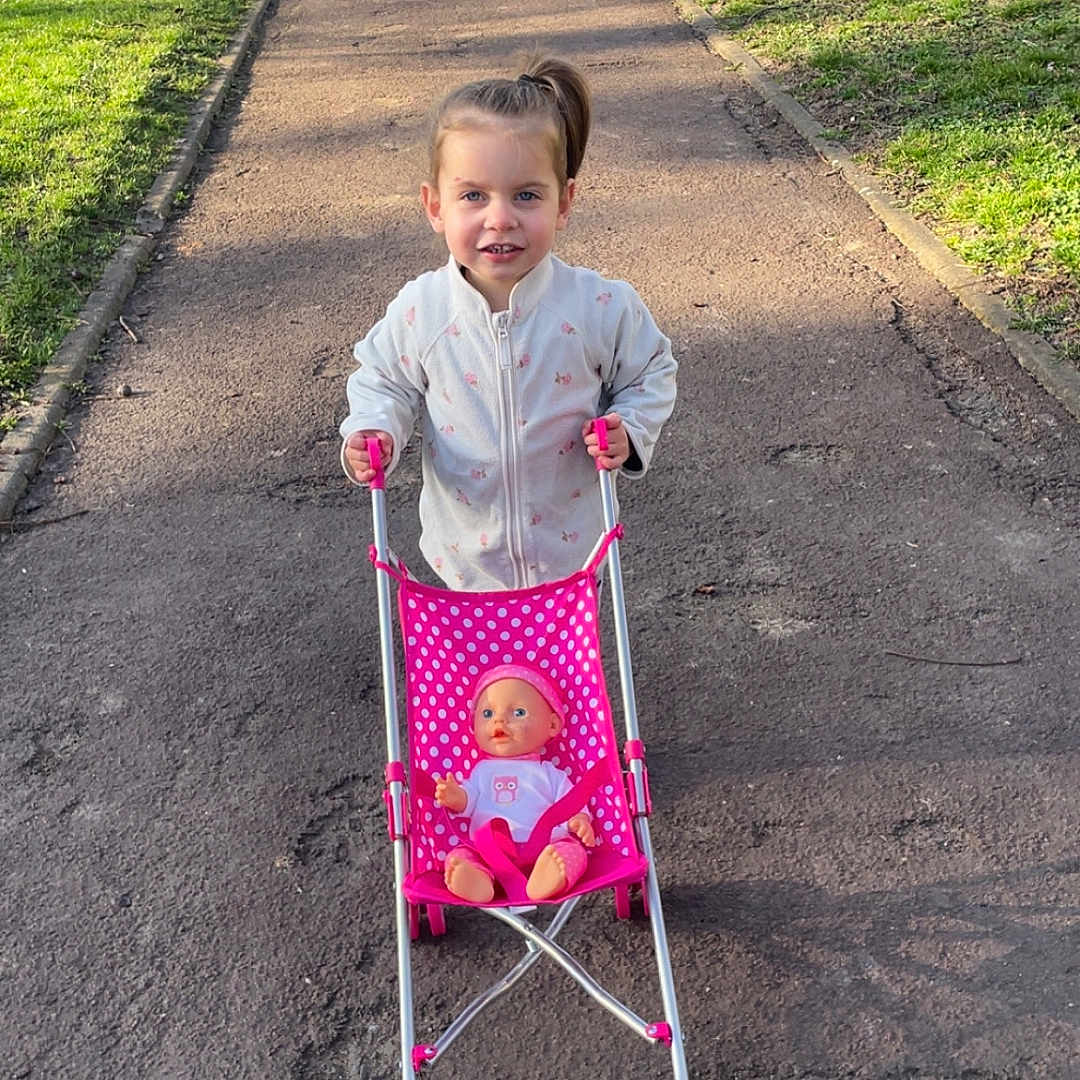 Mya participe au concours pour gagner de l'argent avec cette photo : child, doll, stroller, pink, park, pathway, grass, trees, outdoor, sunny, casual_clothing, smiling, play, toy, person, young_child, daylight, nature, recreation, happy