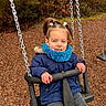 child, swing, playground, blue_jacket, scarf, pigtails, smiling, outdoor, wood_chips, trees, park, play, happy, person, chain, seat, casual_clothing, fall, fun, young_girl