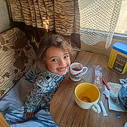 Aelis a rejoint le concours — aidez-le/la à gagner de superbes lots ! blanket, child, clothing, cozy, curtain, drink, face, food, hair, happy, hot_chocolate, indoor, mug, natural_light, person, smiling, snack, table, window, wood