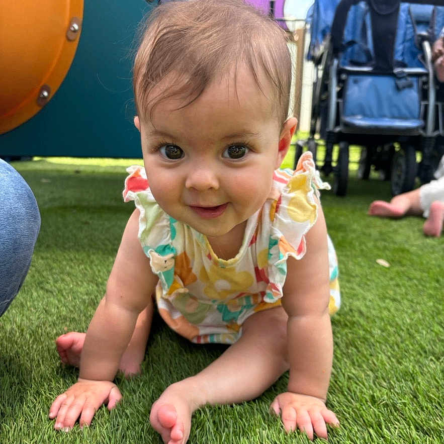 Hollie is registered to the contest to win money with this photo: baby, child, crawling, grass, playground, outdoor, colorful_clothing, stroller, toy, infant, smiling, person, cute, young_child, sunlight, happy, greenery, feet, hands, playtime