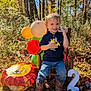 child, toddler, boy, smiling, balloons, toys, wooden_chair, autumn_leaves, grass, outdoor, forest, sunlight, jeans, boots, number_2, celebration, birthday, happy, nature, playful
