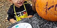 Jaxson is registered to the contest to win money with this photo: child, pumpkin, painting, paint_palette, apron, outdoor, halloween, tablecloth, spiderweb_pattern, smiling, blonde_hair, face, hand, adult, black_hoodie, yellow_apron, craft, fall_leaves, art_supplies, creative