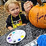 child, pumpkin, painting, paint_palette, apron, outdoor, halloween, tablecloth, spiderweb_pattern, smiling, blonde_hair, face, hand, adult, black_hoodie, yellow_apron, craft, fall_leaves, art_supplies, creative