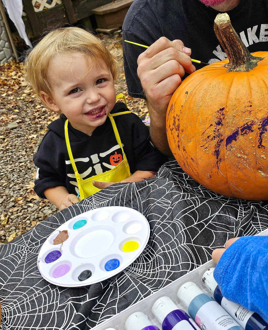 Jaxson is registered to the contest to win money with this photo: child, pumpkin, painting, paint_palette, apron, outdoor, halloween, tablecloth, spiderweb_pattern, smiling, blonde_hair, face, hand, adult, black_hoodie, yellow_apron, craft, fall_leaves, art_supplies, creative