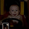 baby, infant, child, smiling, face, portrait, toy_steering_wheel, toy_car, sitting, indoor, home, playroom, dark_lighting, photograph, smartphone_screen, camera_interface, steering_wheel, happy, looking_at_camera, furniture