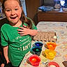 child, green_shirt, smiling, table, colorful_cups, eggs, egg_carton, indoor, decorating, happy, long_hair, wooden_furniture, playful, crafts, celebration, festive, person, home, expression, fun