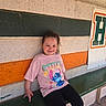 child, girl, sitting, bench, pink_tshirt, stitch, smile, casual_clothing, wall, striped_wall, green_bench, playful, outdoor, daylight, concrete_wall, young_child, happy, short_hair, black_pants, relaxed