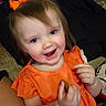 Winnie is registered to the contest to win money with this photo: toddler, child, orange_dress, bow, smiling, happy, face, hand, snack, indoor, person, cute, young_child, hair, sitting, apparel, cheerful, skin, eyes, portrait