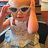 Nova is registered to the contest to win money with this photo: toddler, child, sunglasses, flower_glasses, dress, high_chair, restaurant, table, hair, smile, indoor, person, cute, playful, fashion, accessory, blond, seat, portrait, fun
